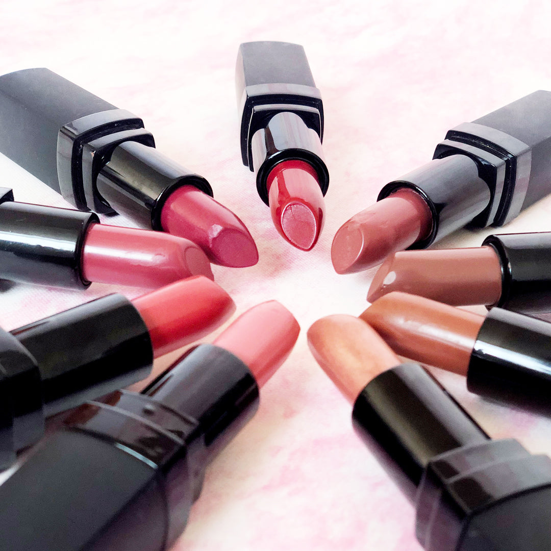 Collection of open lipsticks in various shades arranged in a circular pattern on a light pink background.