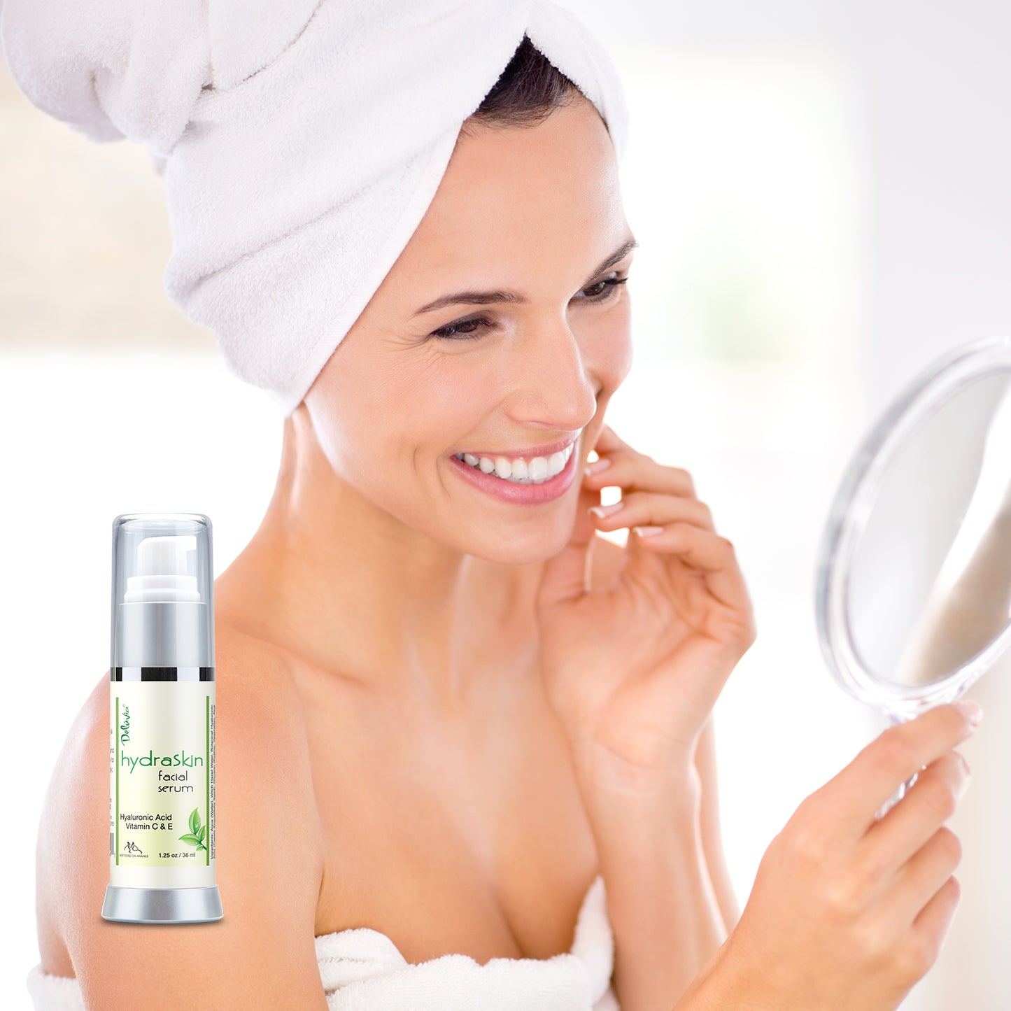A smiling woman touching her face in front of a hand mirror and a Deluvia Hydraskin Facial Serum in the foreground