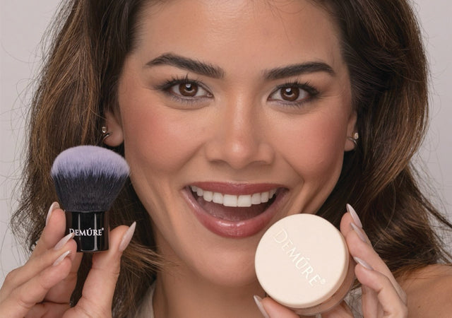Smiling woman wearing Demure Medium Dark Foundation, holding a jar of the foundation and a kabuki brush