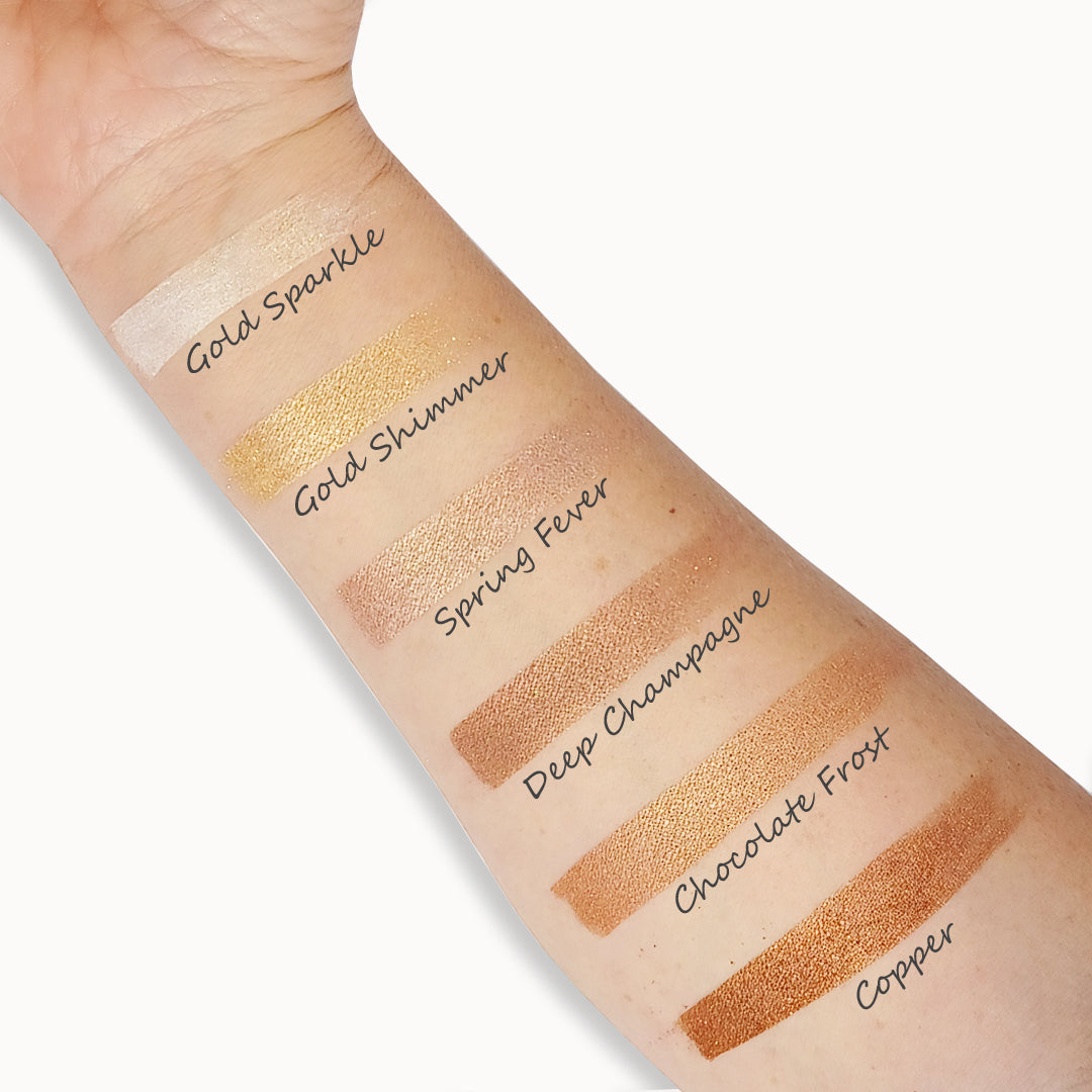 Swatches of different gold and bronze makeup products on a person's arm with labels.