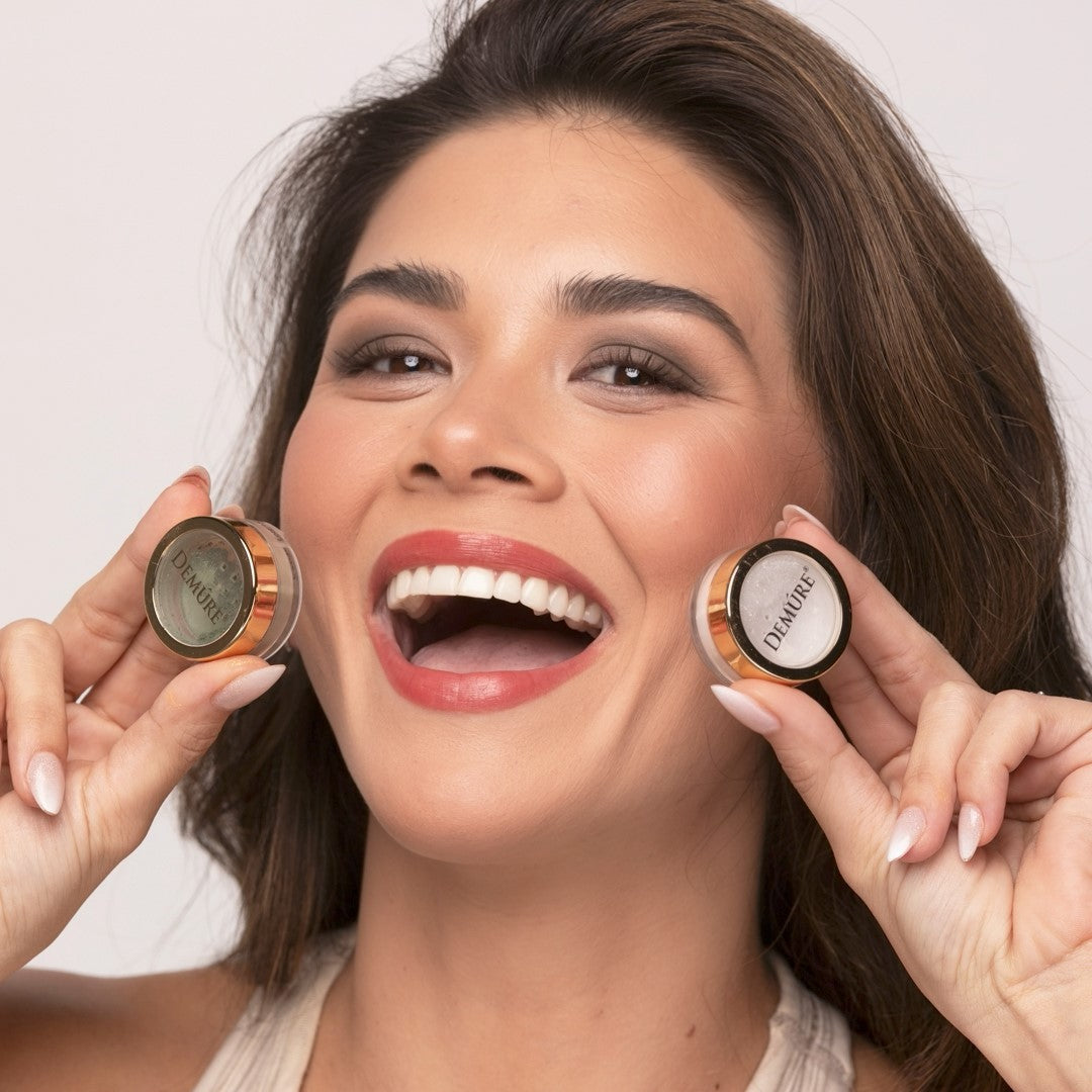 Woman holding two makeup products, Demure Green Khaki eye color and Snowflake eye color close to her face against a plain background