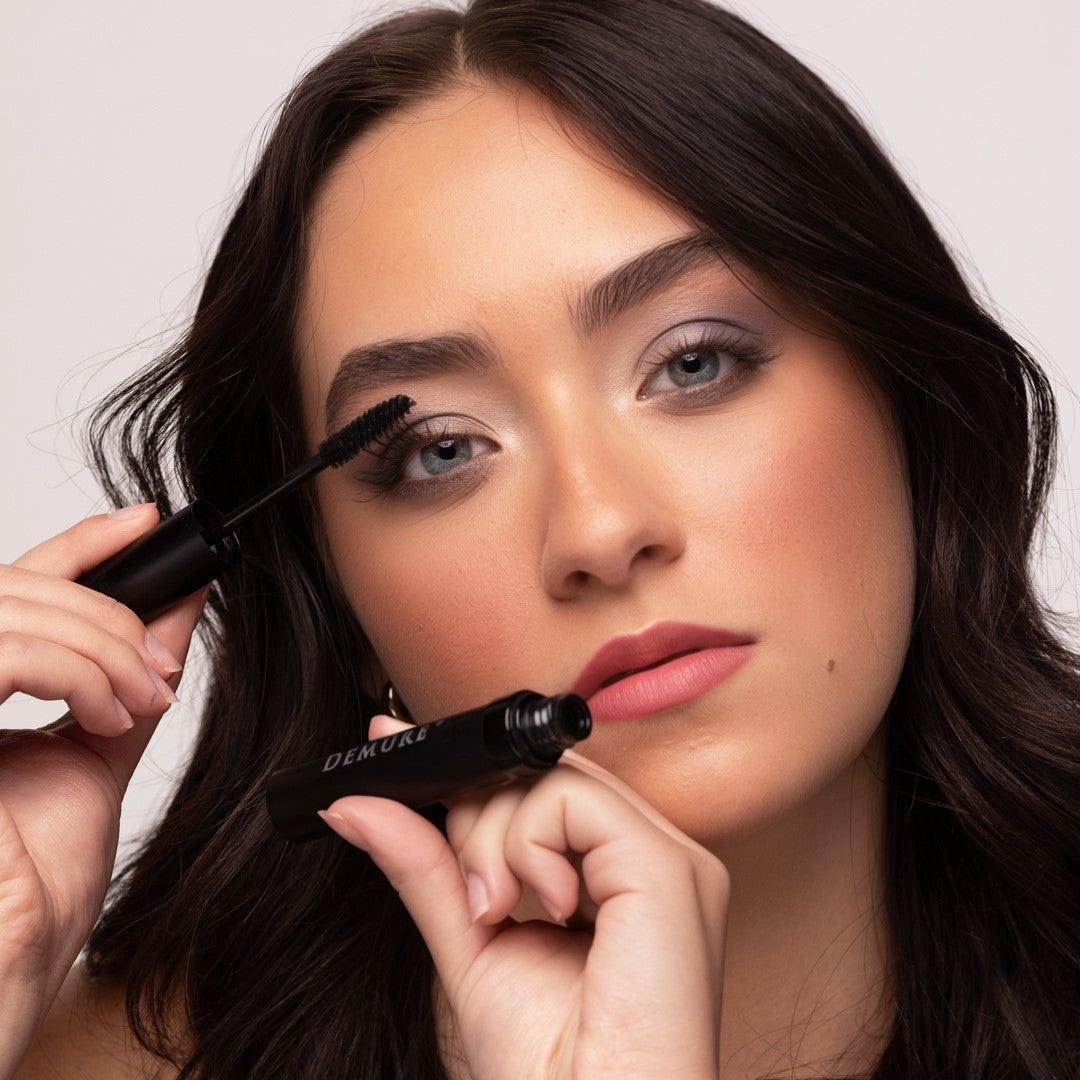 A close-up of a woman applying Demure Black Mascara with one hand while holding the black tube with the other. 