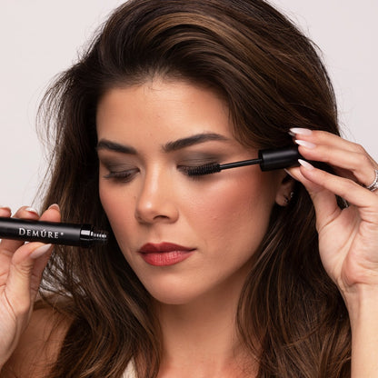 A close-up of a woman applying Demure High Definition Black mascara to her upper lashes, holding the black tube with one hand and the wand with the other, against a light neutral background.