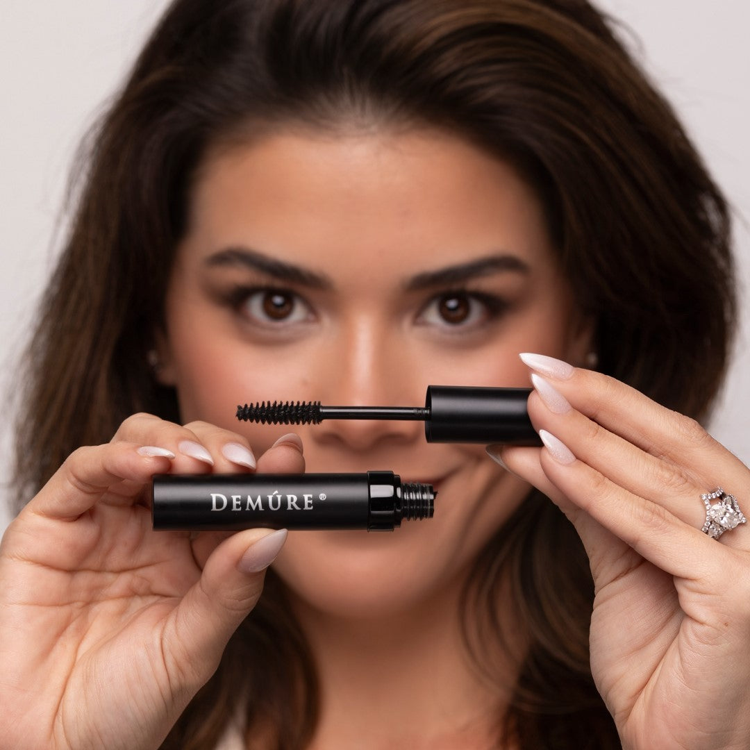 A close-up lifestyle shot of a woman holding a Demure Black Mascara tube and its wand, showcasing the product against her face.