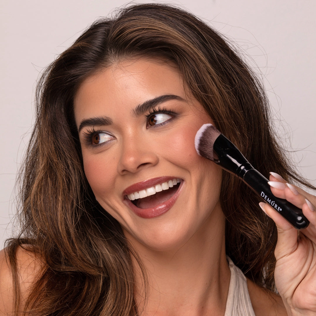 Smiling brunette woman applying Demure Bold Berry Blush to her cheek with a Demure Sculpting Contour Brush