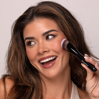 Smiling brunette woman applying Demure Bold Berry Blush to her cheek with a Demure Sculpting Contour Brush
