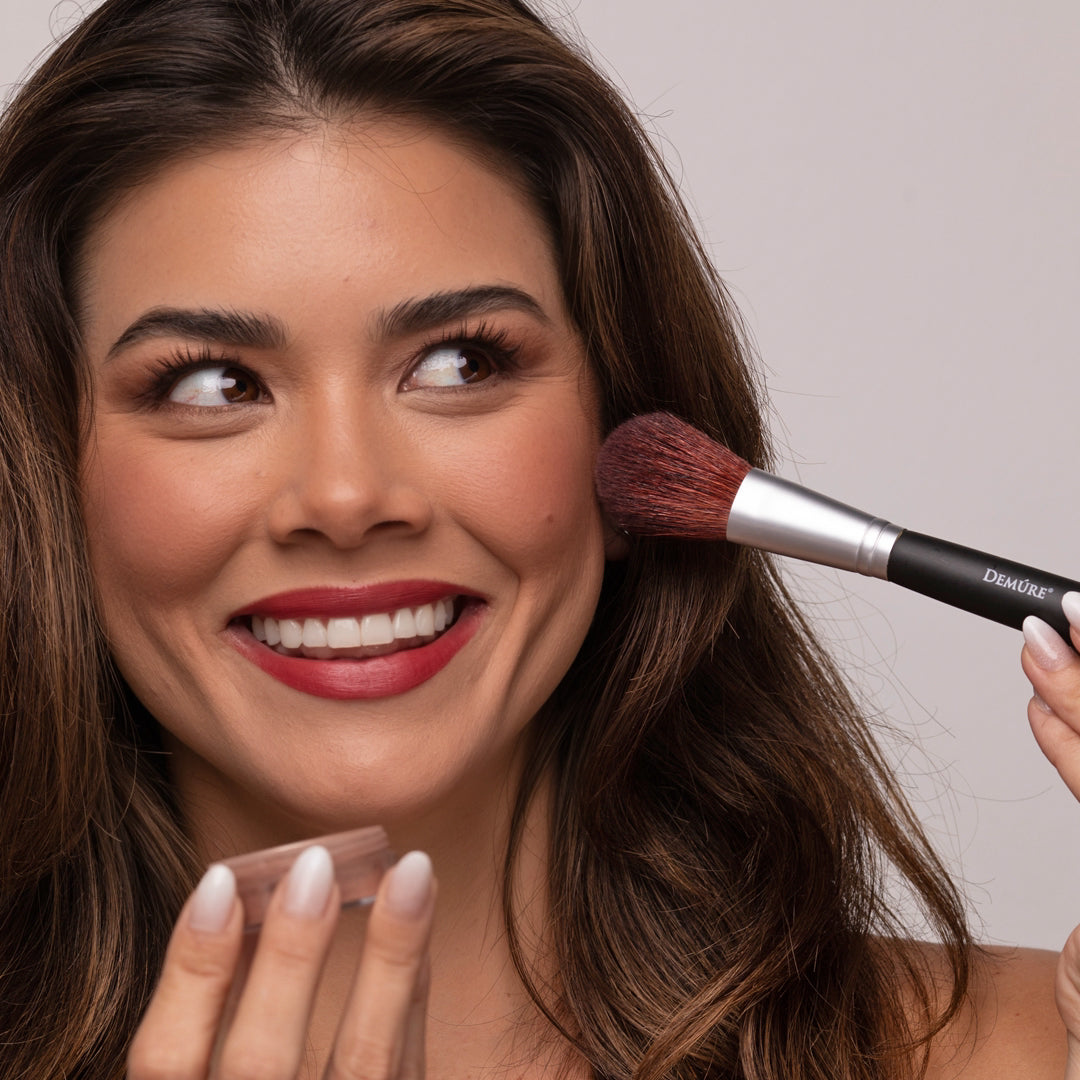 Woman applying Demure Sienna Rose Blush with a brush holding a makeup jar on a neutral background