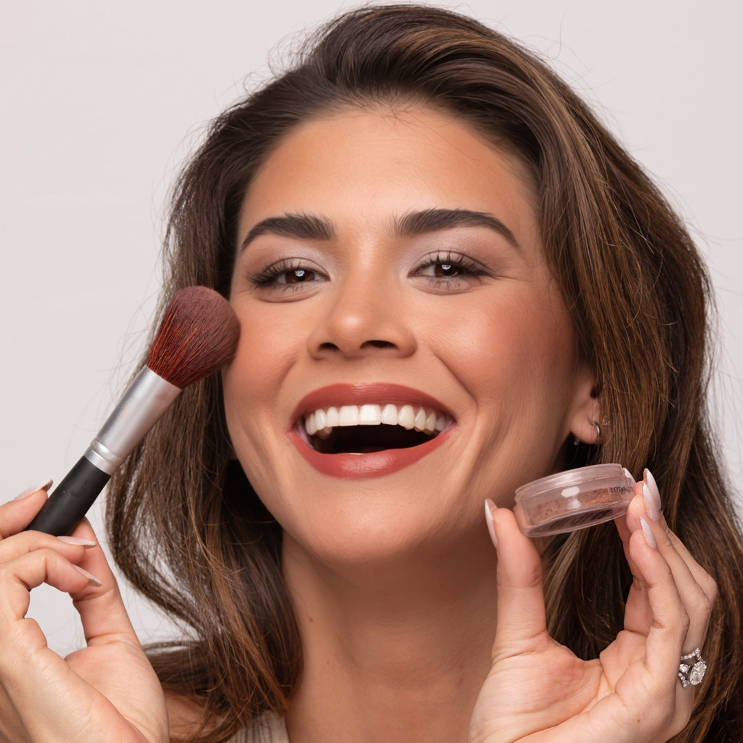 Smiling woman applying Demure Spiced Mocha Blush to her cheek with a Demure Blush Brush, holding a jar of blush color in her hand