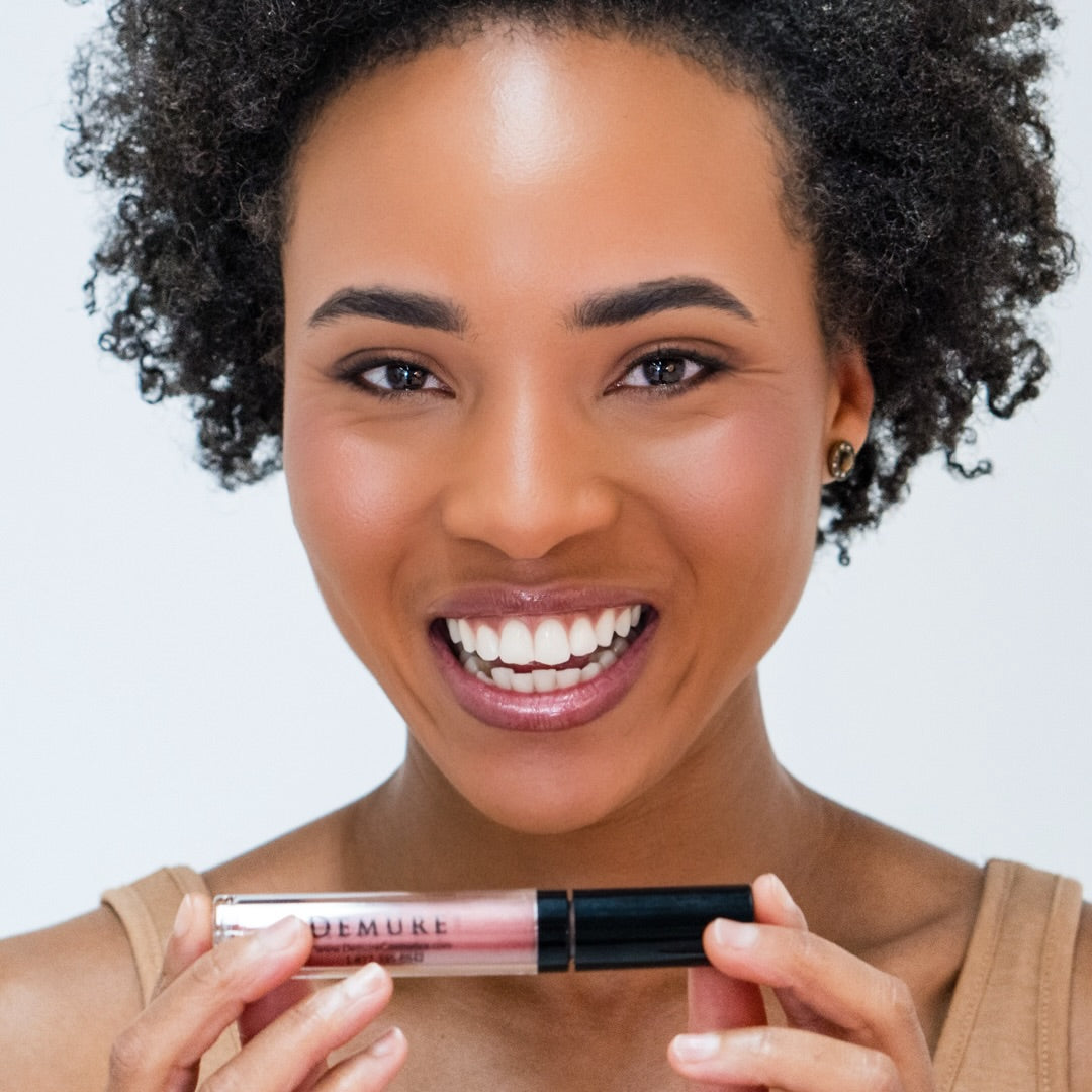 Smiling African American woman holding a tube of Demure Lip Gloss Bronze Glow 30 near her chin, with a white background