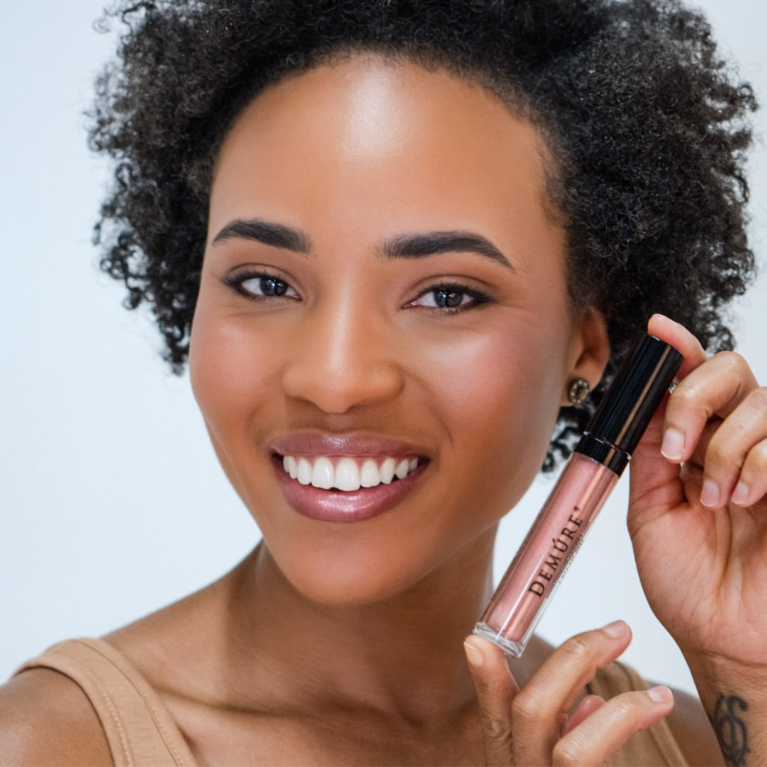 Smiling African American woman holding a tube of Demure Lip Gloss Bronze Glow 30 near cheek, with a white background