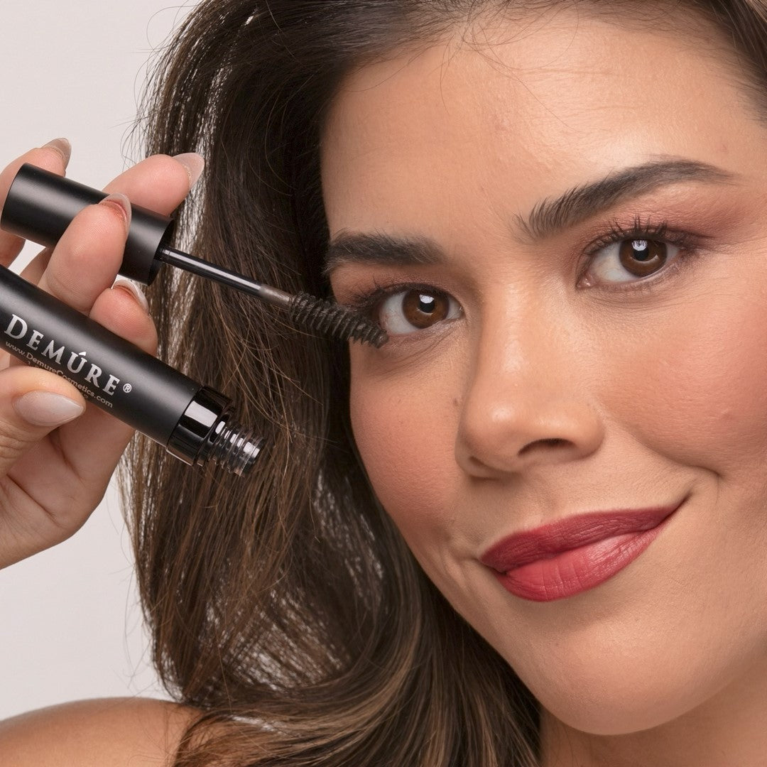 A woman wearing Demure Brown mascara, holding an open tube and mascara applicator