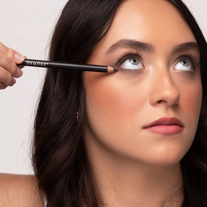 A close-up of a woman looking up, holding a Demure Charcoal eyeliner near her eye.
