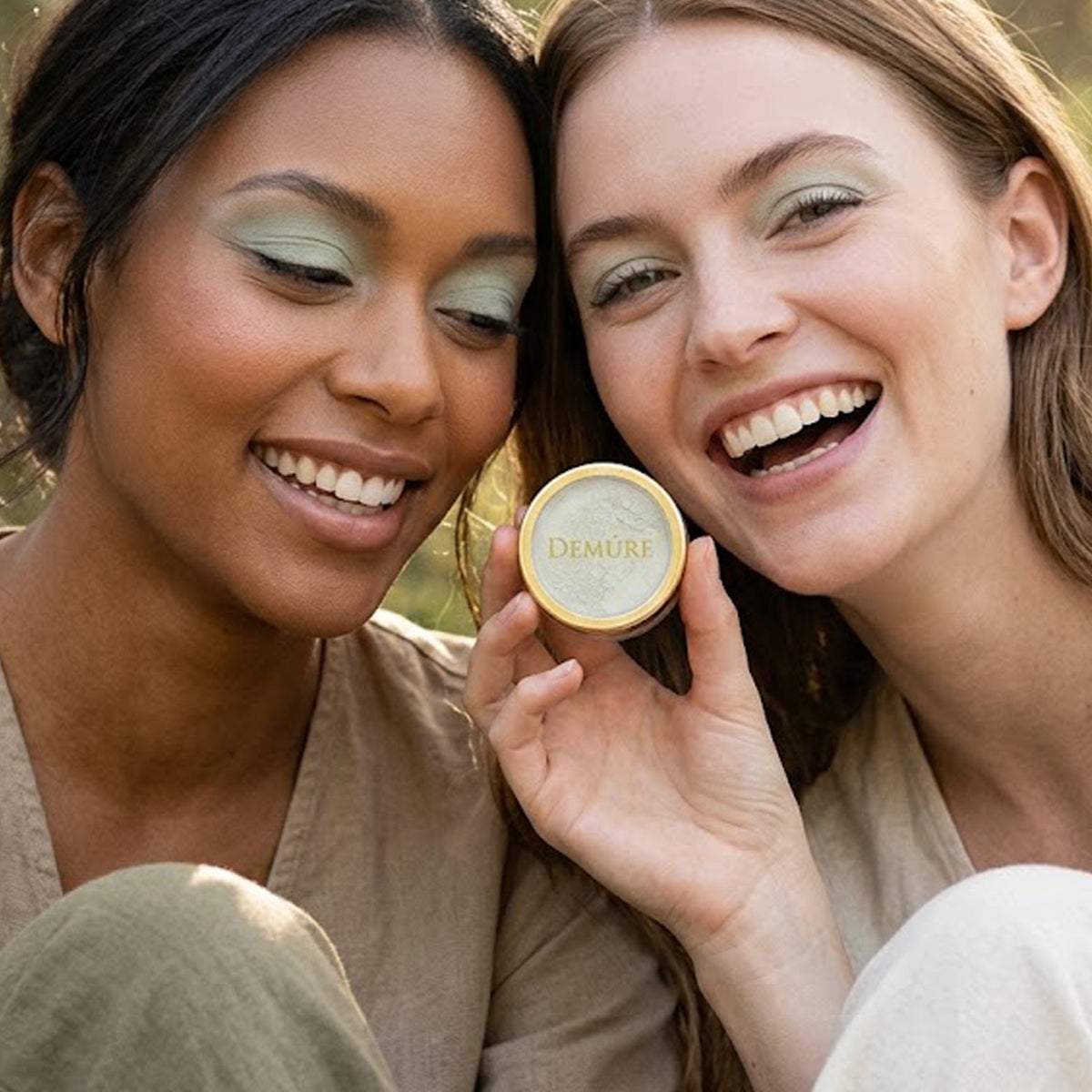 Two smiling women outdoors wearing Demure Coastal Sage eye color, holding a round gold tin of Demure makeup, with one woman showing the product and the other looking on closely.
