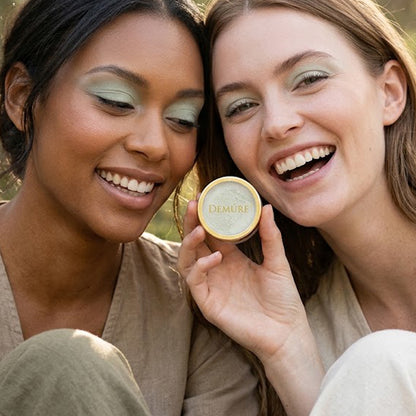 Two smiling women outdoors wearing Demure Coastal Sage eye color, holding a round gold tin of Demure makeup, with one woman showing the product and the other looking on closely.
