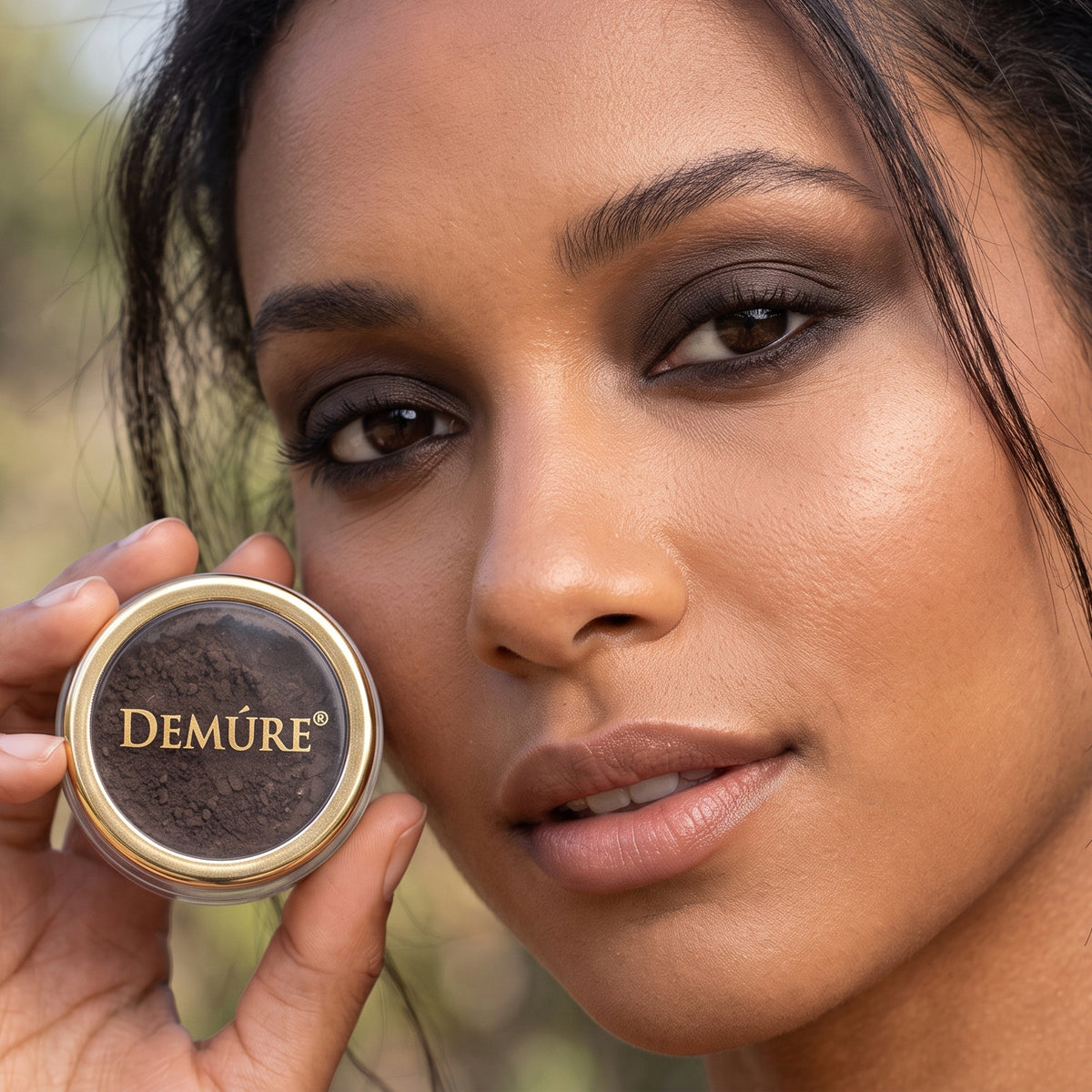 A close-up of a woman holding a Demure eye color Cool Espresso powder makeup next to her face, showing the dark brown product inside.