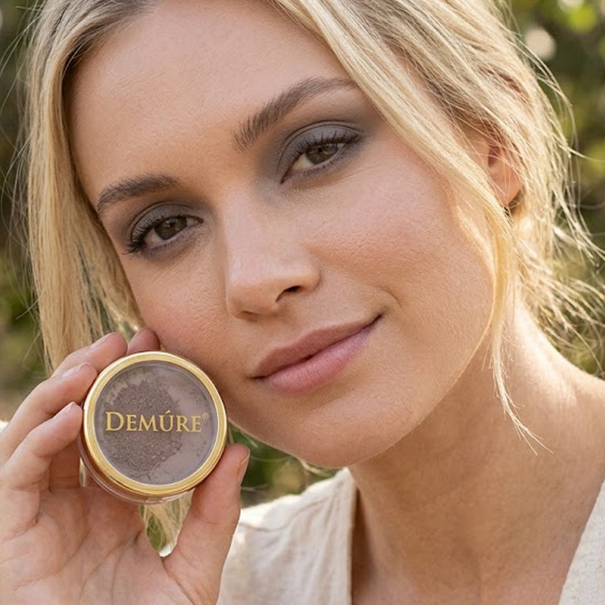 A close-up lifestyle shot of a woman holding a round gold-lidded compact of Demure Driftwood eye color powder next to her face, with a soft-focus outdoor background.