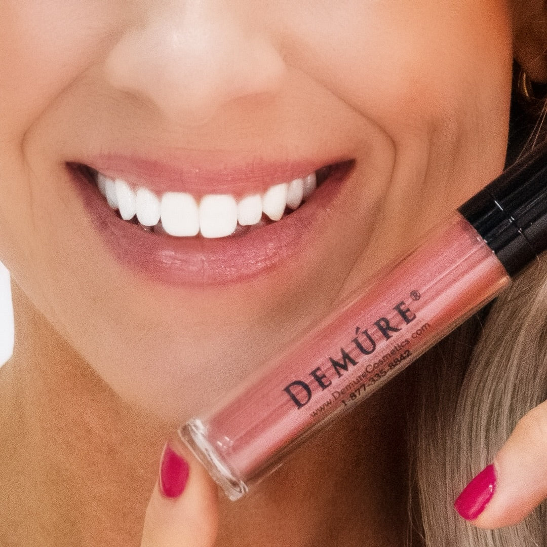 Close up of a woman's lips wearing Demure Lip Gloss Dusty Rose and Demure Lip Liner Antique Rose and holding a tube of the gloss near her lips.