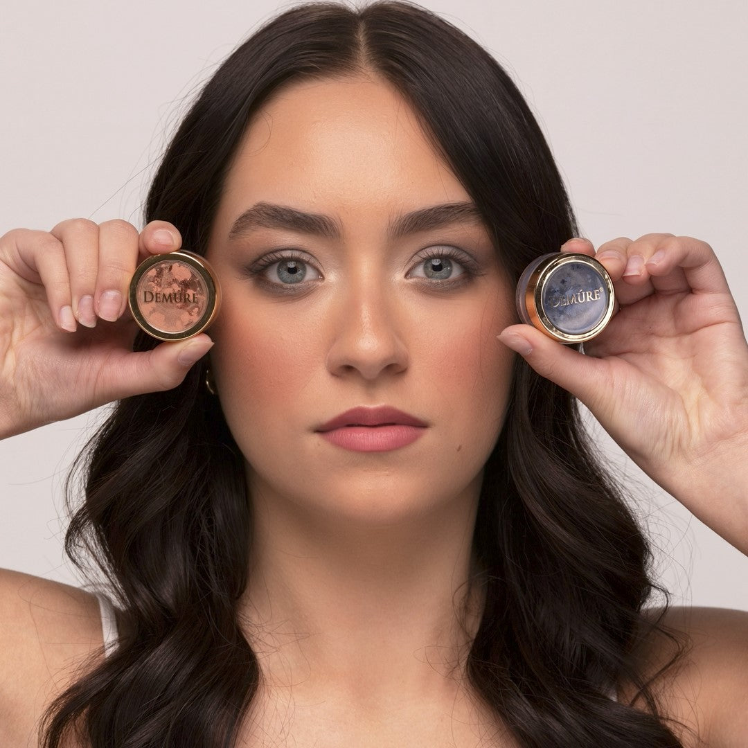 Woman wearing Demure Coral eye color and Blue Sparkle eye color and holding the jars near her eyes.
