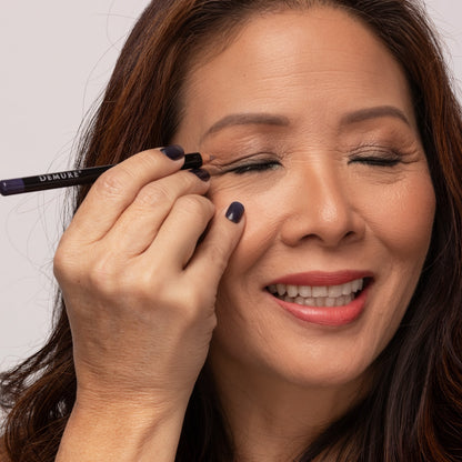 A close-up of a woman with closed eyes as she applies Demure Charcoal eyeliner.