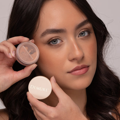 Woman holding an open jar of Blush Go Bronze near her cheek and wearing Go Bronze Blush