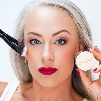 Woman applying Demure Go Bronze Blush with a contouring brush and jar 