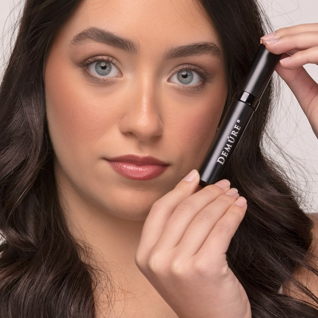 Woman holding a tube of Demure High Definition Black Mascara near her eyes.