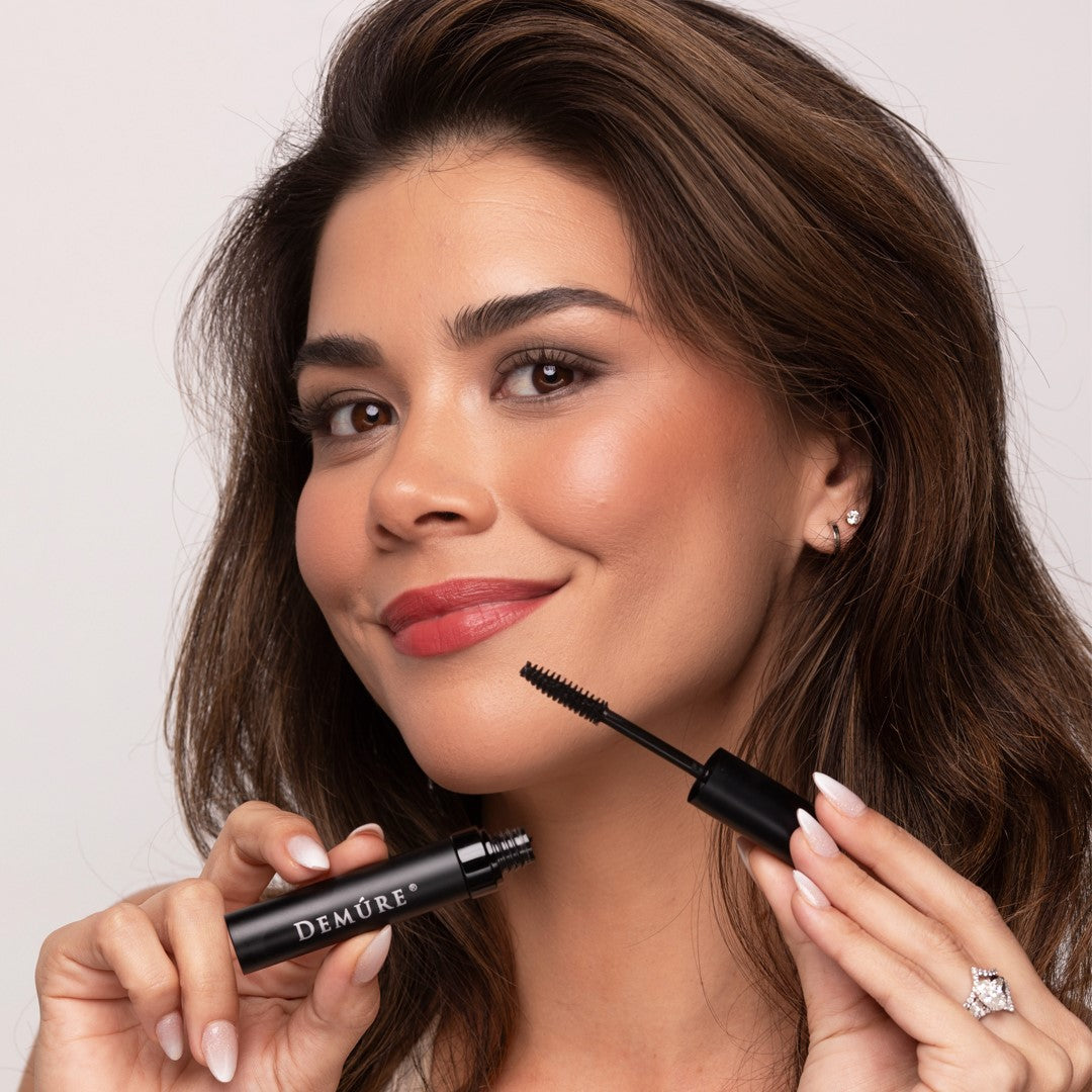 A woman holds a High Definition Black Demure mascara tube and its wand near her face against a light neutral background.