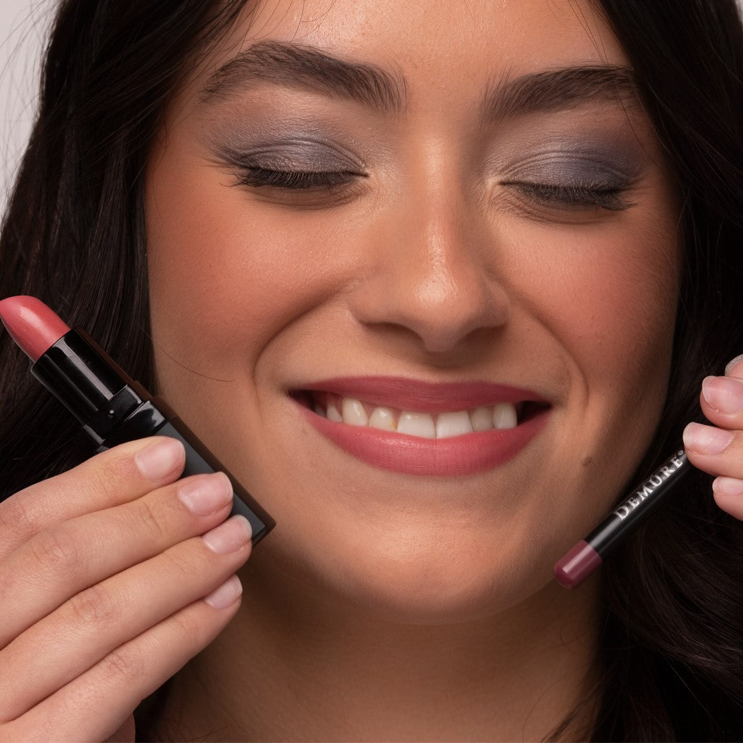 A close-up of a woman with closed eyes wearing Demure Soft Rose lipstick and holding it in one hand and a matching lip liner pencil in the other, both shown against her face.