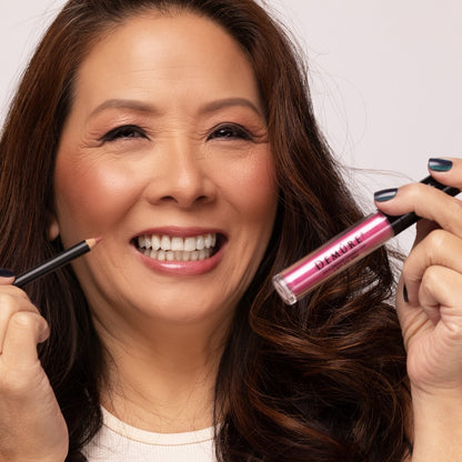 A smiling woman with long brown hair holds a Demure Lilac Shimmer lip gloss tube in one hand and a Demure Le Rose Lip liner pencil in the other, set against a clean light gray background.