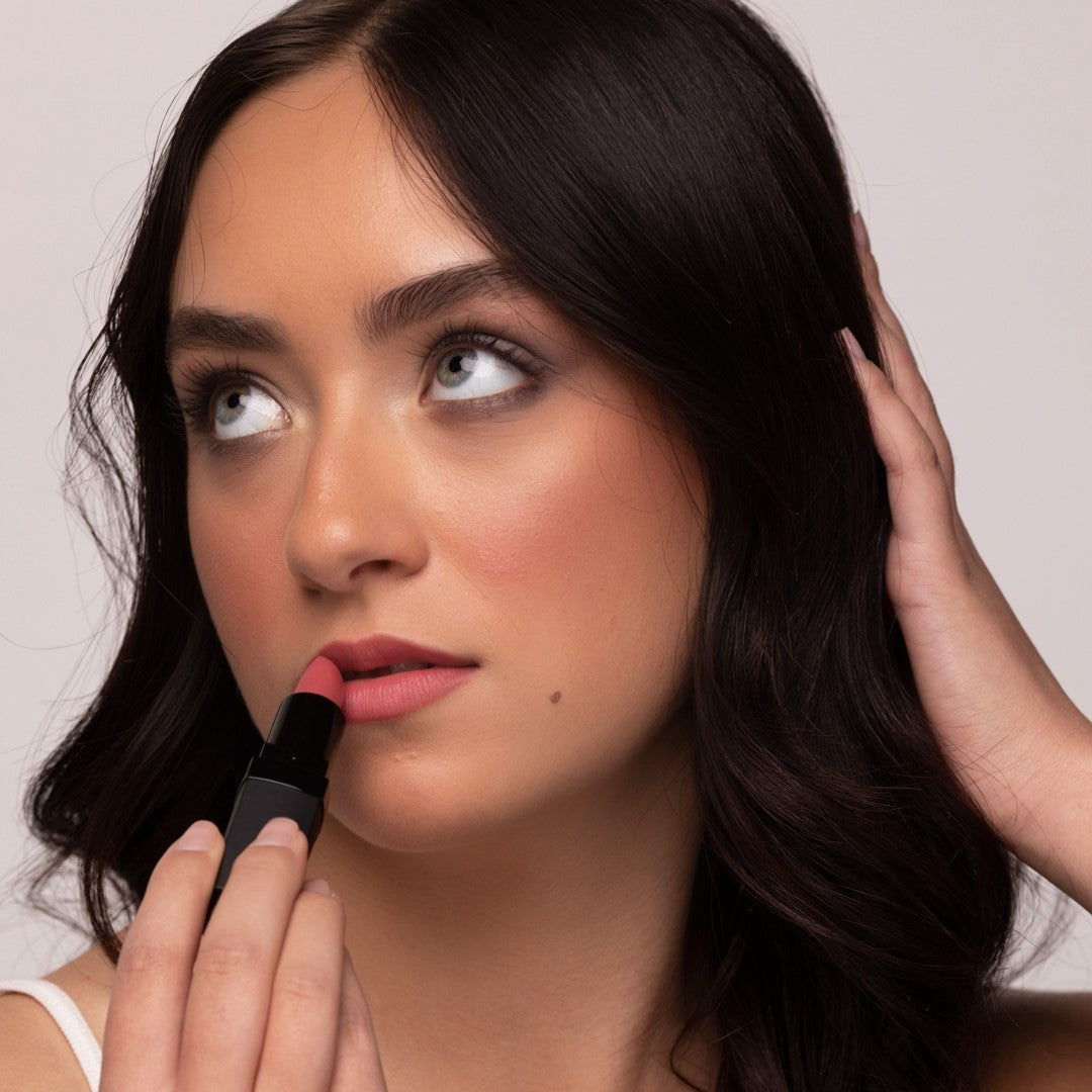 A close-up of a woman with dark hair applying Demure Soft Rose lipstick to her lips against a light neutral background.