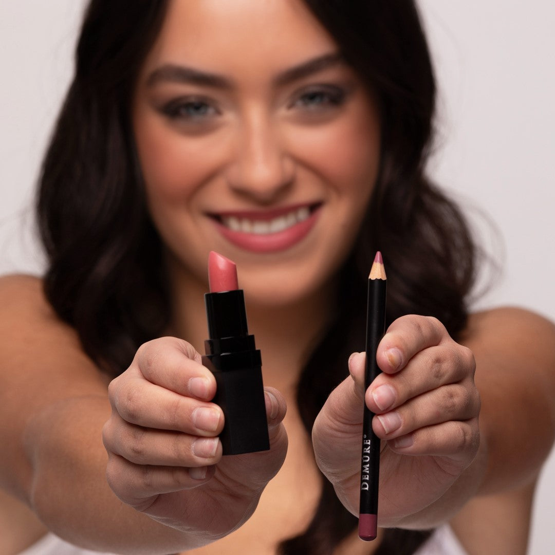 A smiling woman holds a Demure Soft Rose lipstick and a matching Antique Rose  lip pencil toward the camera. 