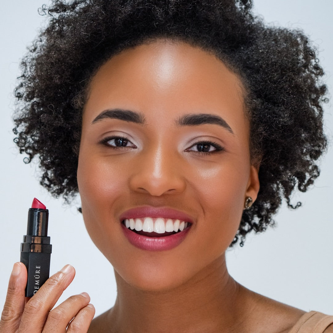 Woman wearing Demure Sweetheart Lipstick 698 and holding an open lipstick with a plain background
