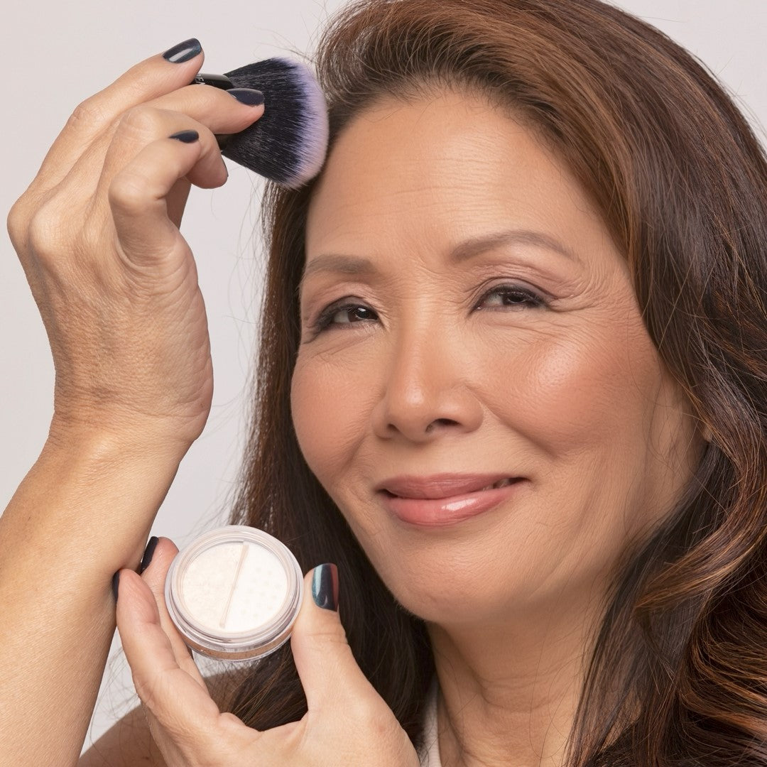 Woman applying Demure Medium Dark Warm Foundation with a Kabuki brush, holding a jar of foundation.