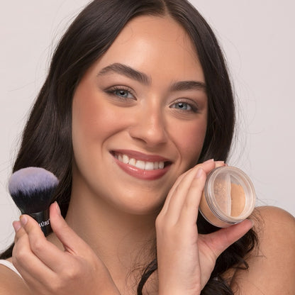 Woman holding a makeup brush and container of Demure Medium Warm Foundation powder against a neutral background