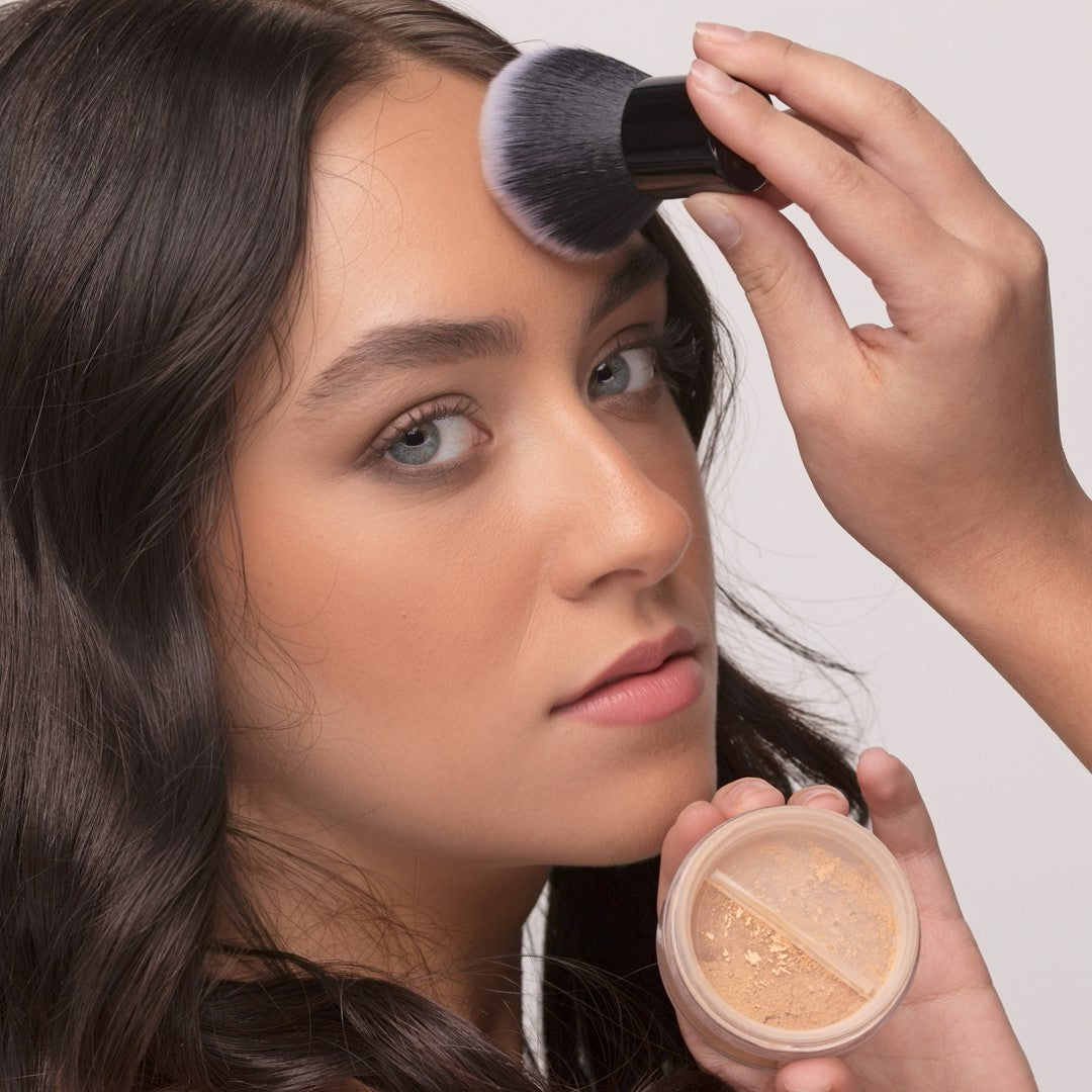 Woman using Kabuki Brush to apply foundation and holding a jar of makeup