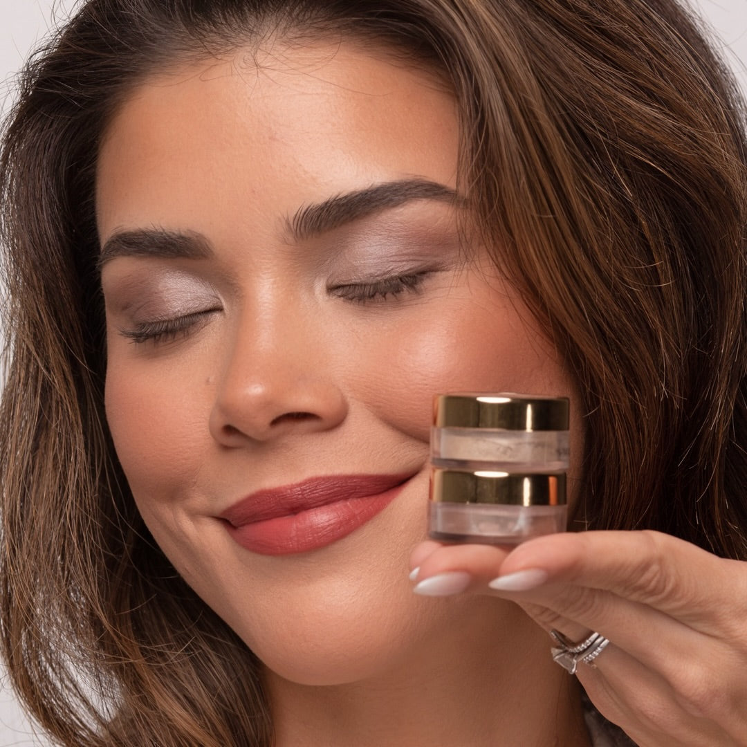Woman with her eyes closed wearing Demure eye colors Mocha Ice and Dusty Rose, holding the two jars in her hand