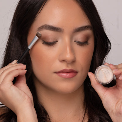 Woman wearing Demure Naked Glow and Copper eye colors, applying with a  brush and holding an open jar