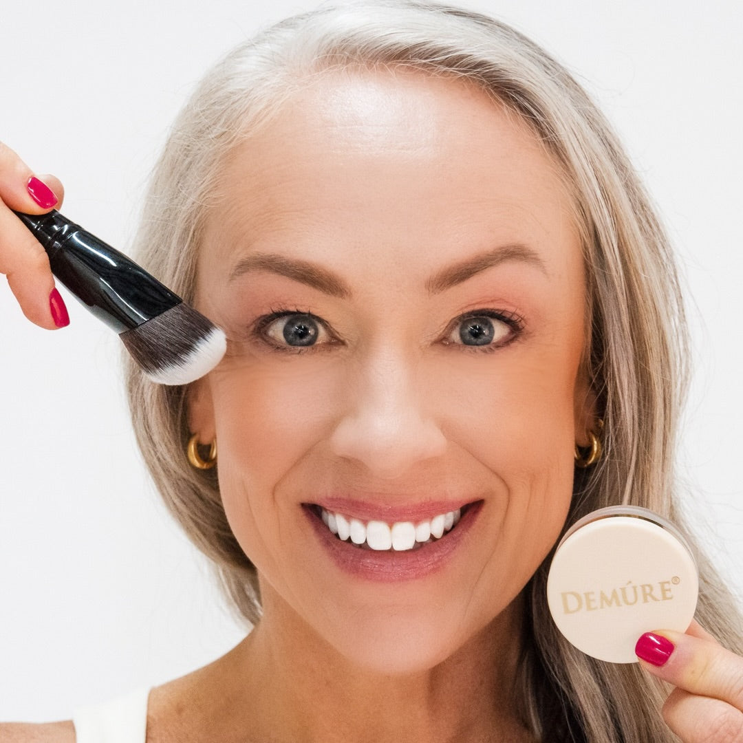 Woman wearing Demure Natural Radiance Blush holding a jar of blush near her right cheek and a contour brush by her left cheek