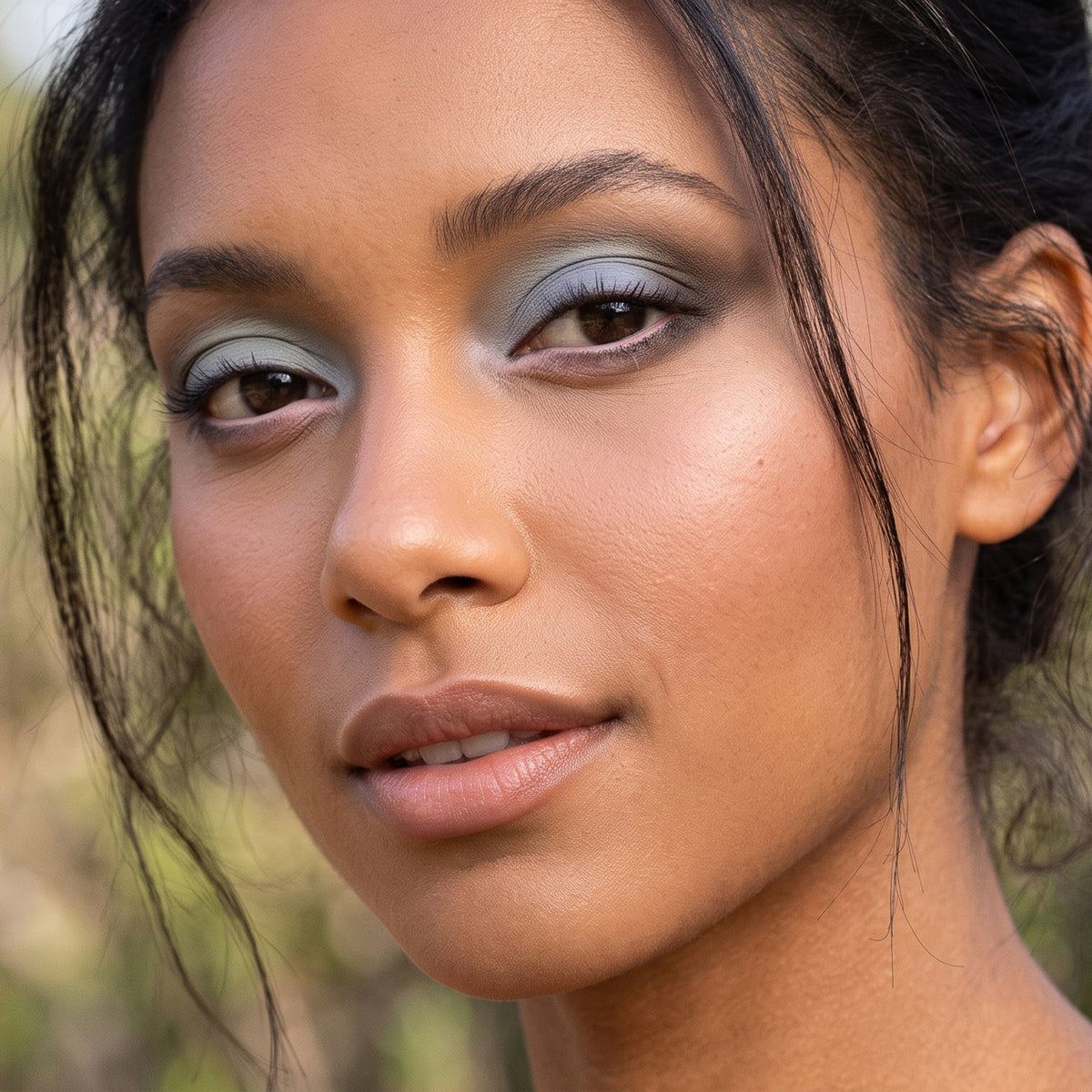 Close-up portrait of a woman wearing Demure eye colors, smoky eyeshadow in muted blue-gray tones, with warm-toned skin and dark hair against a softly blurred outdoor background.