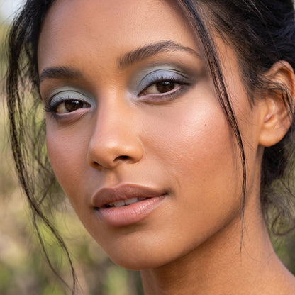 Close-up portrait of a woman wearing Demure eye colors, smoky eyeshadow in muted blue-gray tones, with warm-toned skin and dark hair against a softly blurred outdoor background.