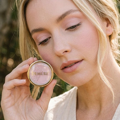 A close-up of a woman holding a gold round jar of Demure Orchid Pink eye color, and wearing the color on her eyes, showing the light pink shimmer product inside, with a soft-focus outdoor background.