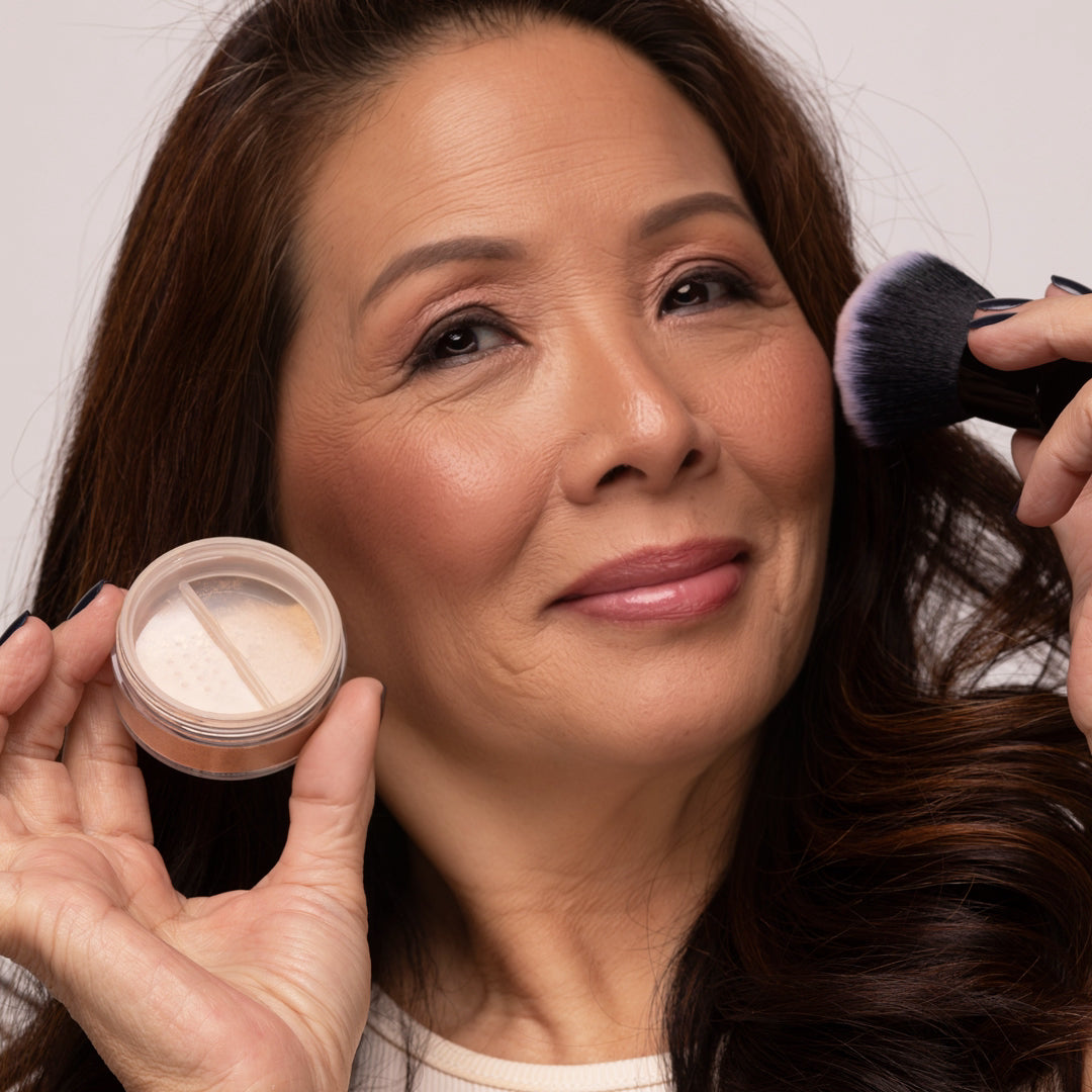 Woman applying Demure Pink Fusion Blush with a brush and container, against a neutral background