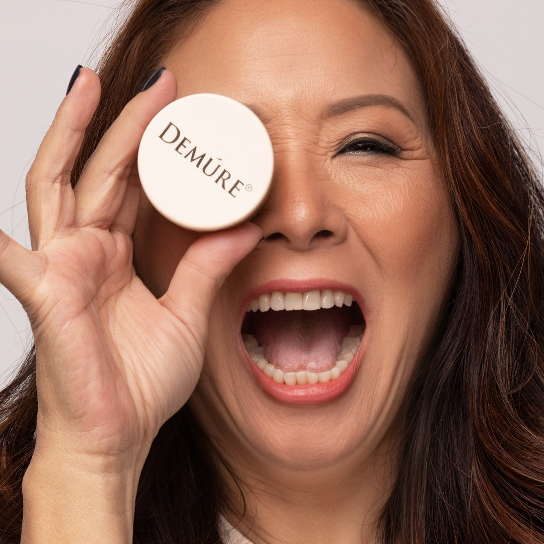 A close-up of a woman holding jar of Demure Tan Finishing Powder in front of one eye, with her other eye closed in a playful wink. The background is a clean, light off-white studio backdrop.