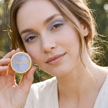 A woman outdoors wearing Demure Washed Denim eye color, showing a light lavender-blue pressed powder. She has matching light purple eyeshadow on her eyes.