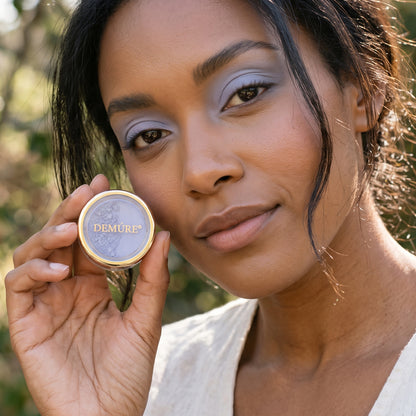 A close-up of a woman outdoors wearing Demure Washed Denim eye color, holding a jar. She has matching light purple eyeshadow on her eyes.