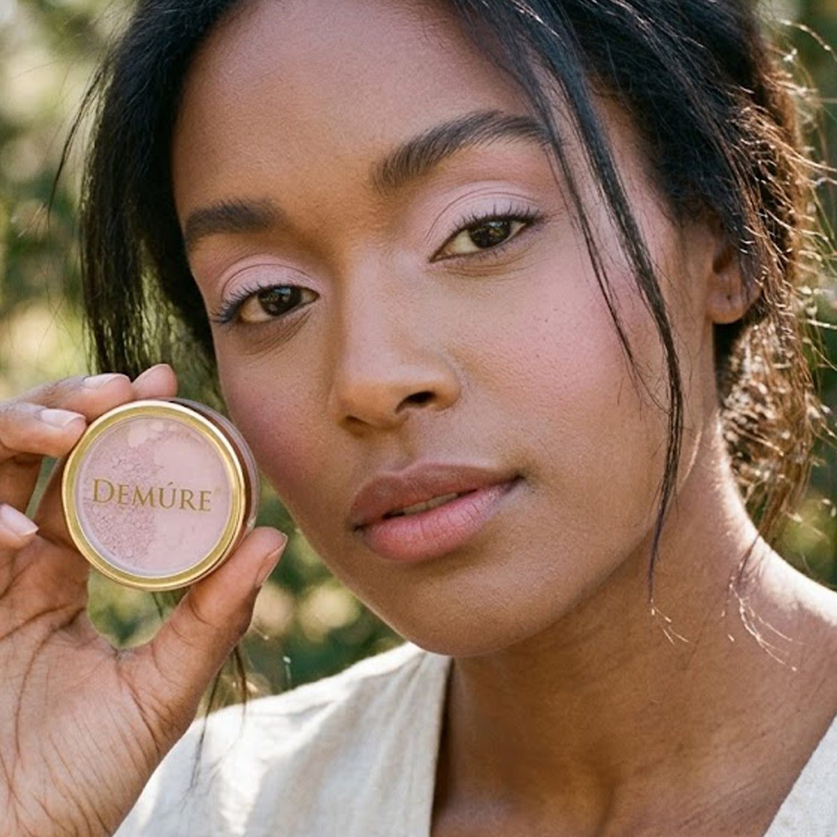 A close-up lifestyle shot of a woman wearing Demure Orchid Pink eye color, holding a jar next to her cheek, showcasing the light pink product against a soft, blurred outdoor background.