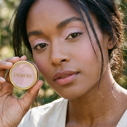 A close-up lifestyle shot of a woman wearing Demure Orchid Pink eye color, holding a jar next to her cheek, showcasing the light pink product against a soft, blurred outdoor background.