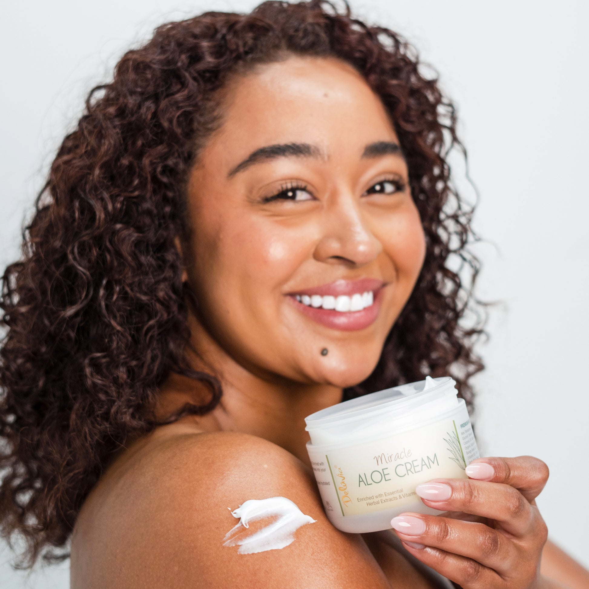 Smiling African American woman holding an open Deluvia Miracle Aloe Cream with some cream on her shoulder