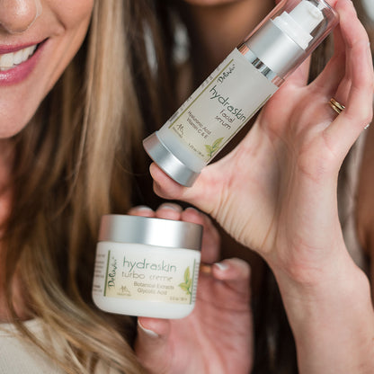 Two women holding jars of Deluvia Hydraskin Turbo Creme and a pump of HydraSkin Facial Serum