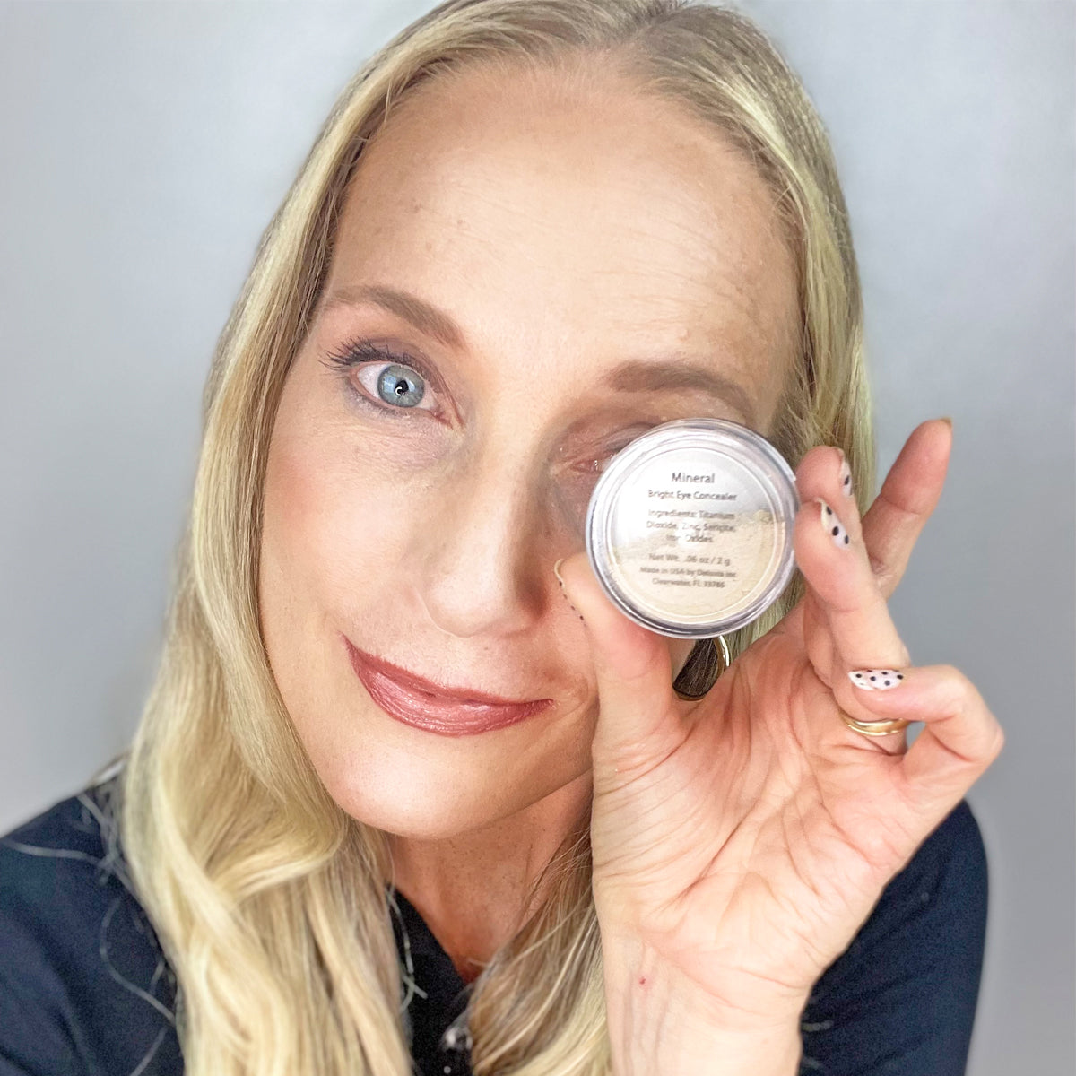 A woman holding a jar of the concealer next to her eye.